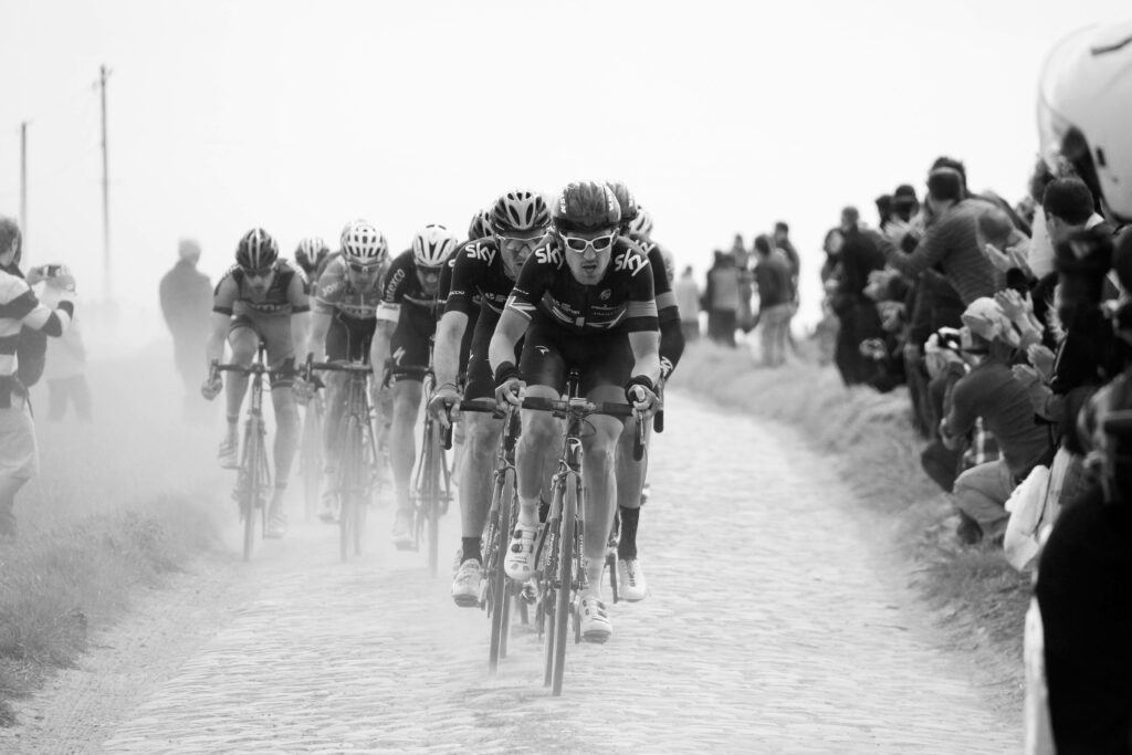 Intense cyclists race on sandy cobblestone road with crowds and dynamic dusty atmosphere grayscale Wallpaper