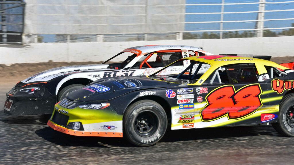 Two colorful stock cars racing side by side on sunlit dirt track showcasing vibrant paint schemes Wallpaper