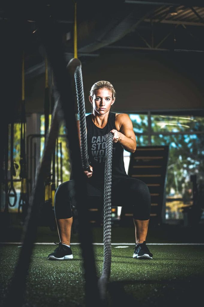 Focused female athlete doing deep squat with battle ropes in moody professional gym wallpaper background