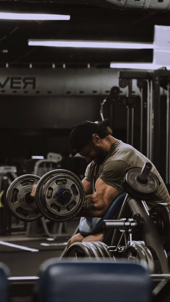 Focused bearded man doing barbell bicep curl in dim industrial gym high-contrast workout wallpaper