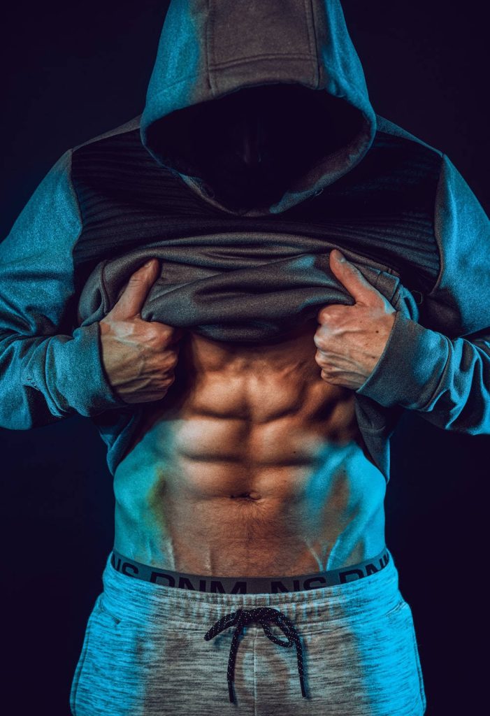 Muscular bodybuilder lifting grey hoodie showing six-pack abs in moody blue lighting fitness wallpaper