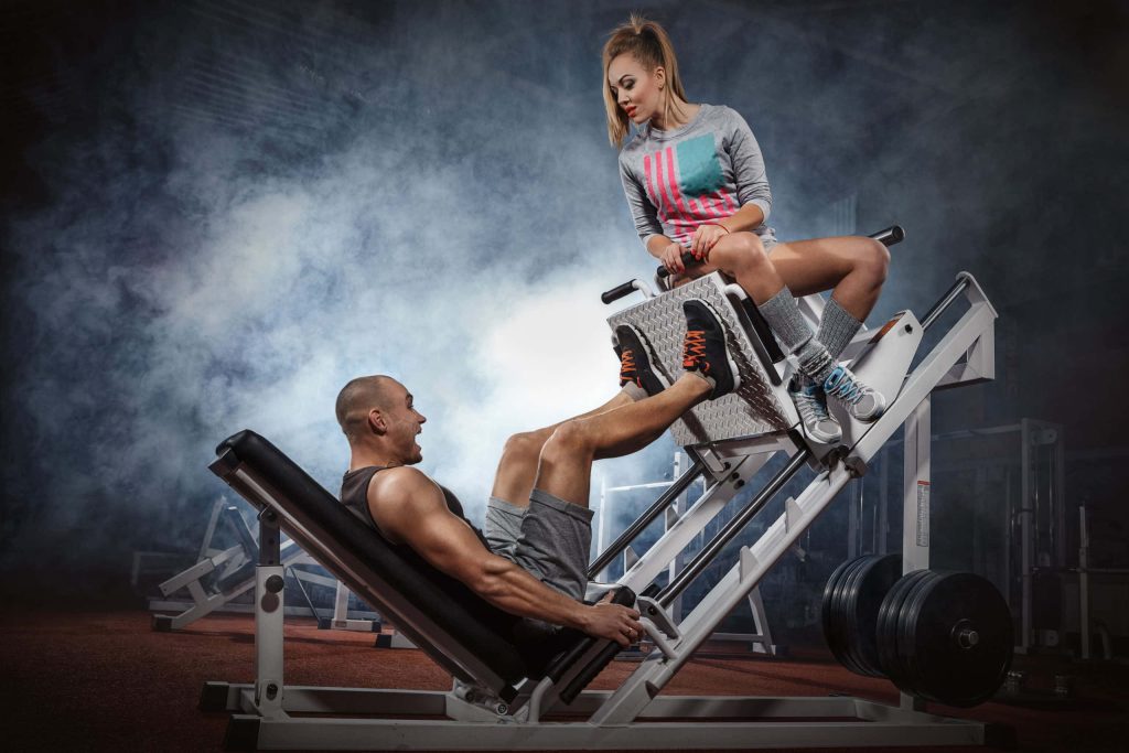 Muscular man doing leg press workout in smoky modern gym supervised by woman trainer Wallpaper