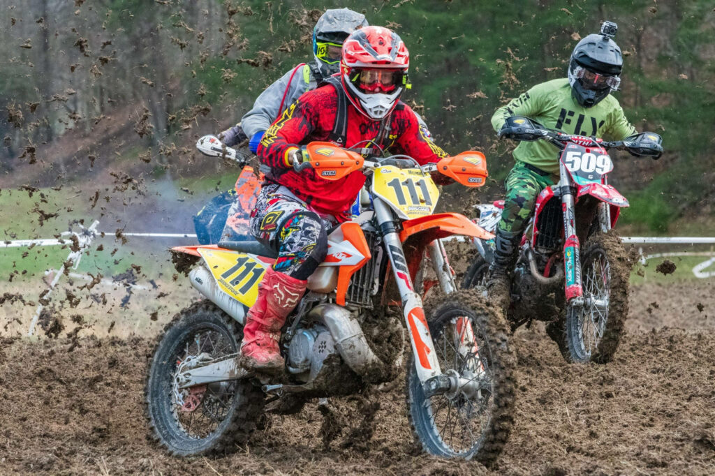 Three motocross riders in colorful gear race on muddy off-road track surrounded by greenery Wallpaper