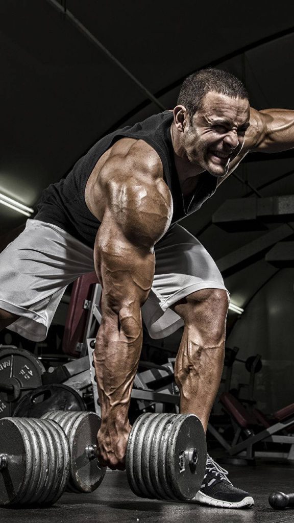 Muscular bodybuilder lifting heavy dumbbell in dramatic gym with intense gritty lighting Wallpaper