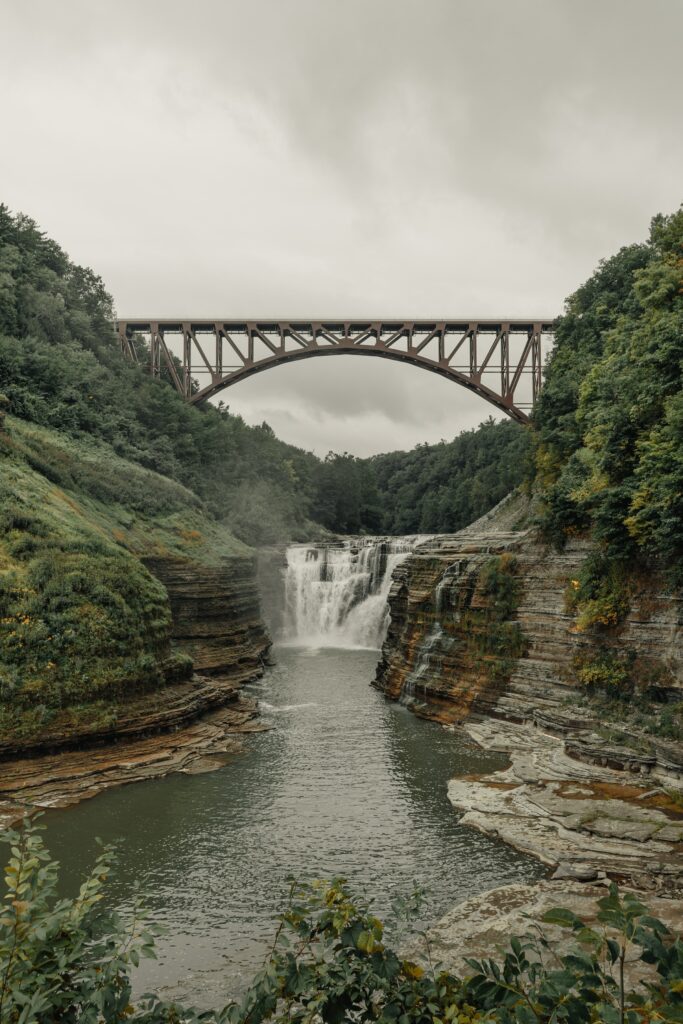 Tall iron bridge over waterfall with lush green forest and cloudy sky serene landscape wallpaper