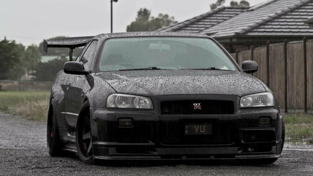 Black Nissan Skyline GT-R R34 with aggressive JDM stance and custom GT wing in rain Wallpaper
