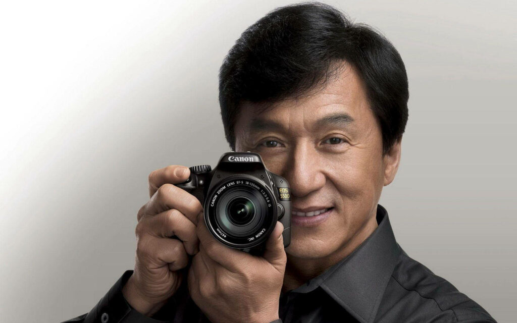Jackie Chan smiling holding Canon camera close-up vibrant wallpaper showcasing photography passion