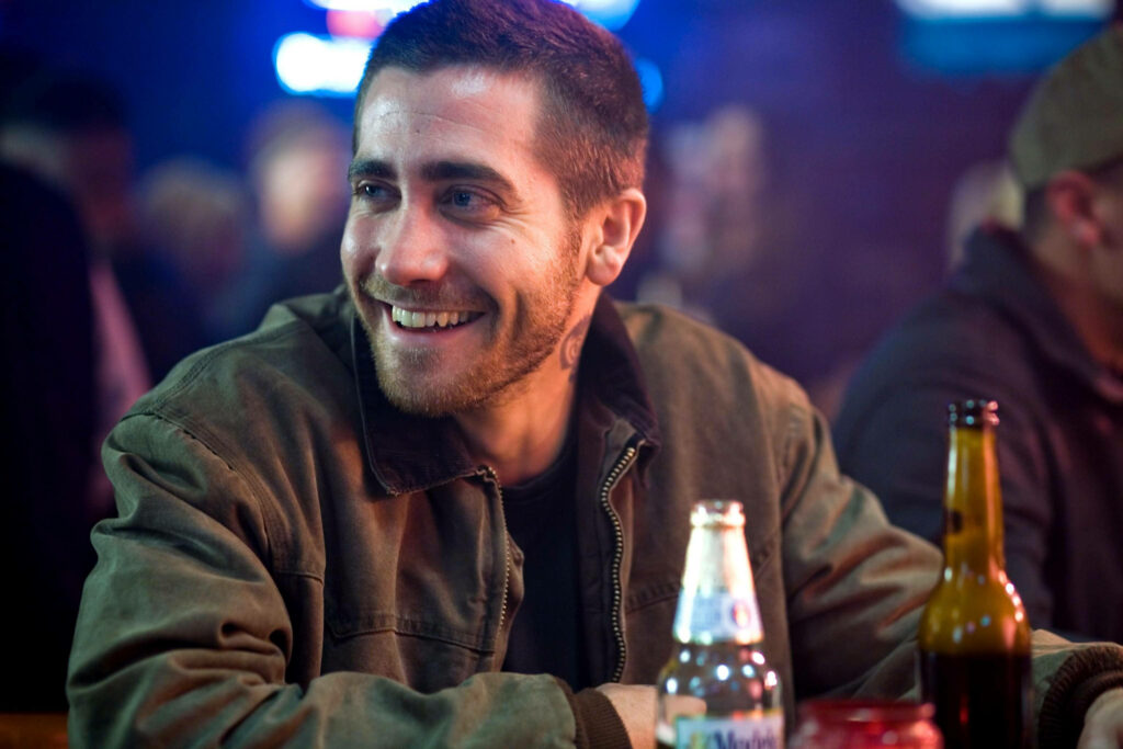 Jake Gyllenhaal smiling warmly in brown jacket at lively bar creating relaxed inviting atmosphere Wallpaper