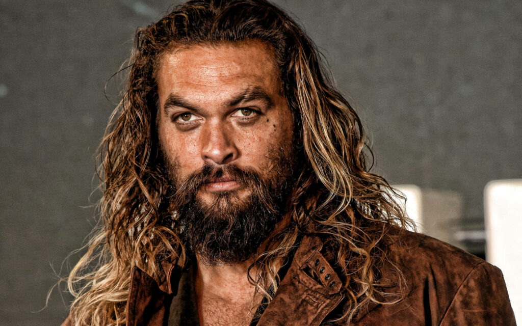 Jason Momoa intense gaze rugged beard long wavy hair brown leather jacket dark background Wallpaper