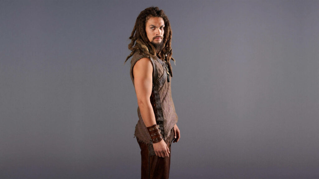 Jason Momoa with iconic dreadlocks and rugged attire stands confidently on grey background wallpaper