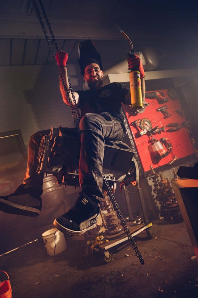 Jaydee from Herencia de Patrones welding in red-lit garage workshop on suspended engine block Wallpaper