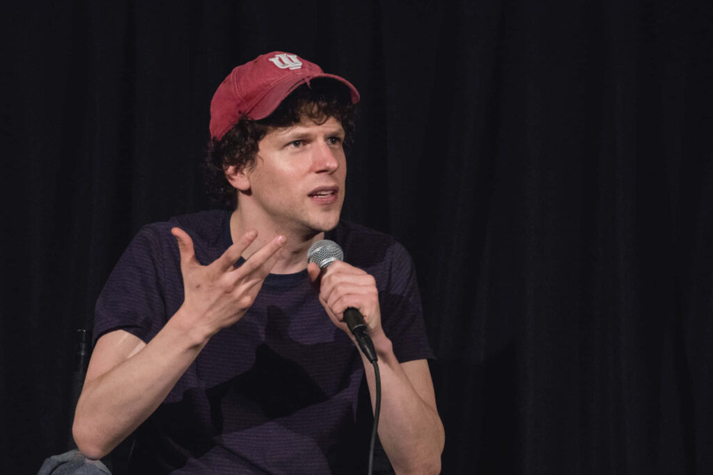Jesse Eisenberg speaking into a microphone wearing red cap dark t-shirt black background wallpaper