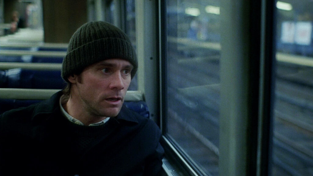 Jim Carrey sitting on train wearing dark beanie and coat gazing thoughtfully out window winter scene Wallpaper