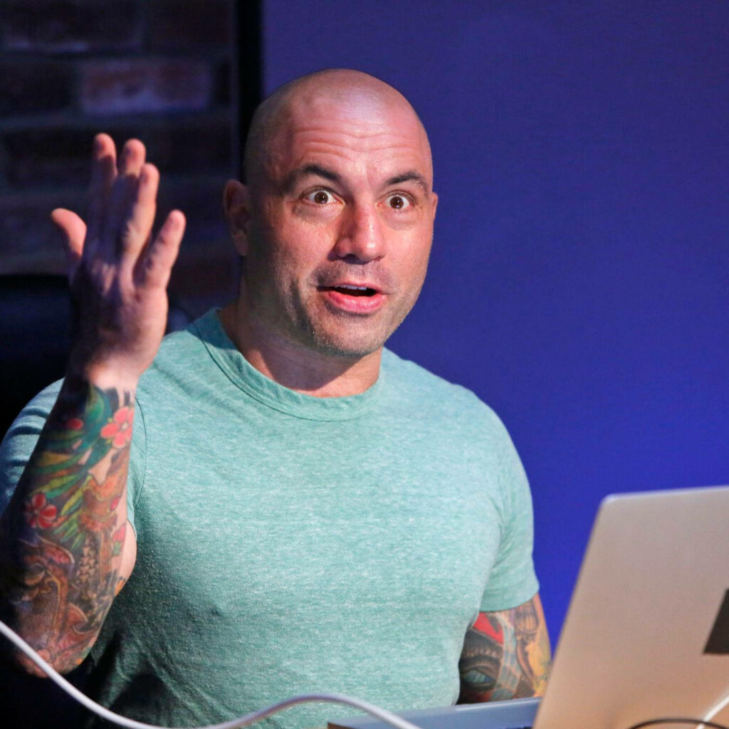 Joe Rogan podcast host animatedly talking on Spotify set with vibrant arm tattoos and laptop Wallpaper