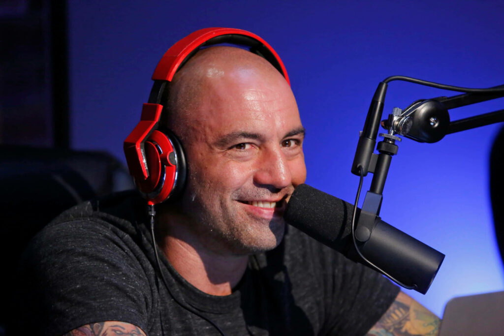 Joe Rogan hosting popular podcast wearing red headphones speaking into microphone blue background Wallpaper