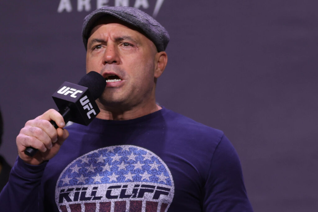 Joe Rogan announces UFC event holding microphone wearing casual hat graphic shirt on wallpaper background