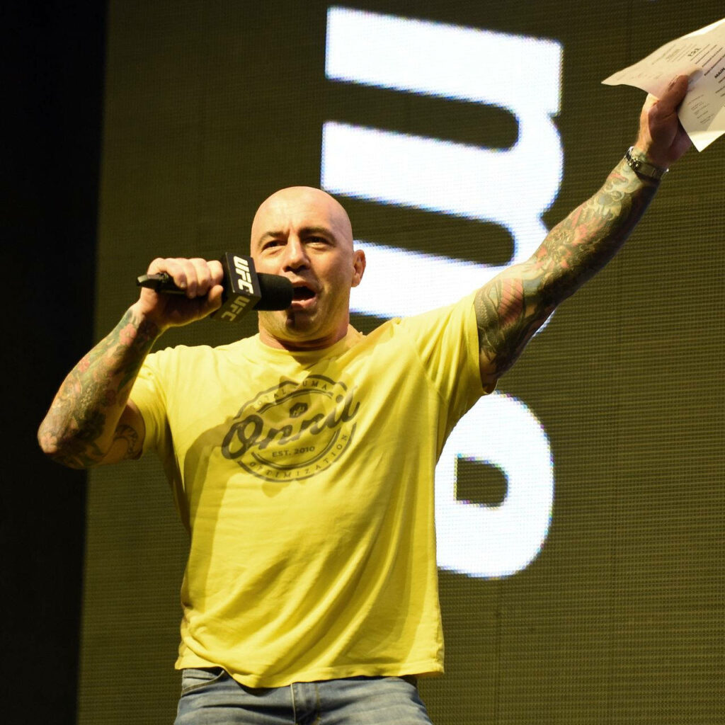 Joe Rogan introducing UFC fighter on stage holding microphone and notes with UFC backdrop wallpaper