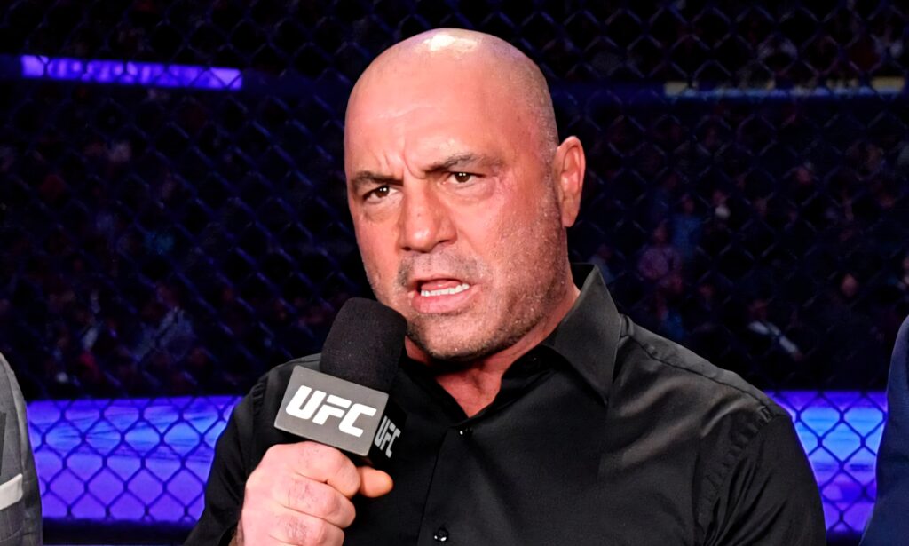 Joe Rogan speaking passionately inside UFC octagon holding microphone with cage and lights background Wallpaper