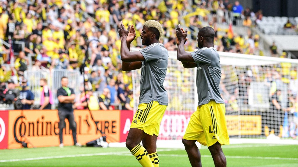 Jonathan Mensah clapping for fans in Columbus Crew kit on pitch with cheering stadium crowd background Wallpaper