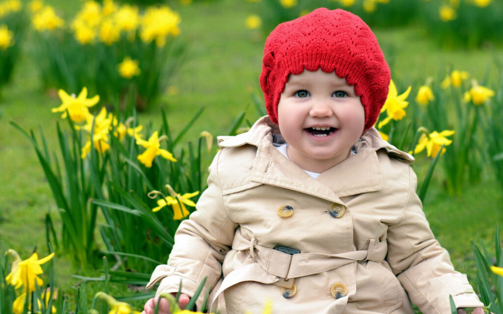 Happy child in beige coat and red hat smiling among yellow daffodils springtime wallpaper background