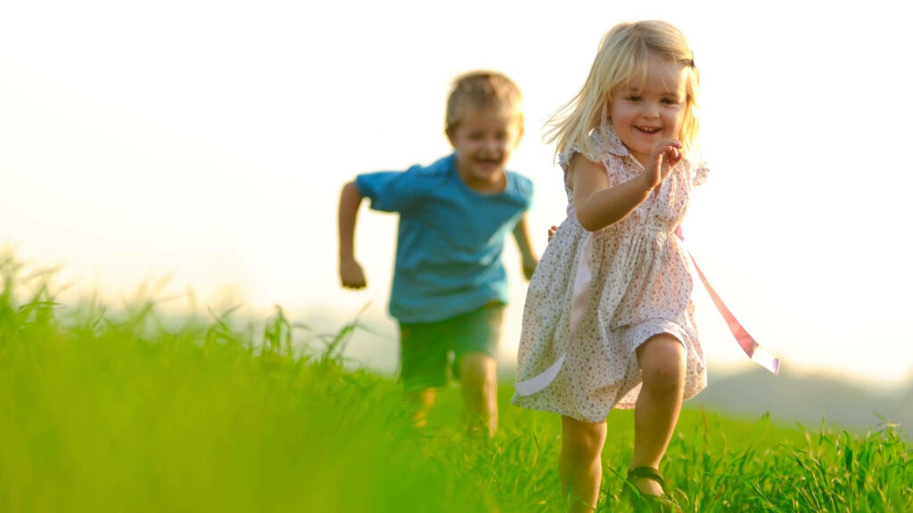 Two happy children running on green grass under bright sky playful innocence wallpaper background