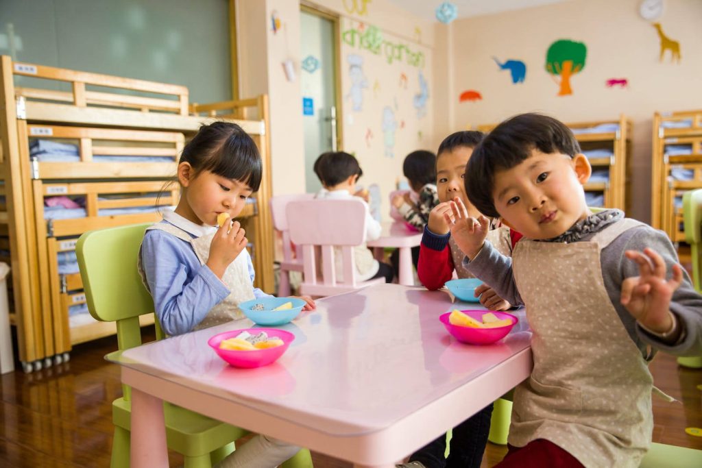 Happy children eating healthy fruit snacks in colorful kindergarten classroom with playful decor Wallpaper