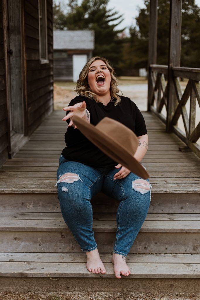 Plus-size joyful woman tossing brown hat on rustic porch steps in casual black tee and jeans Wallpaper