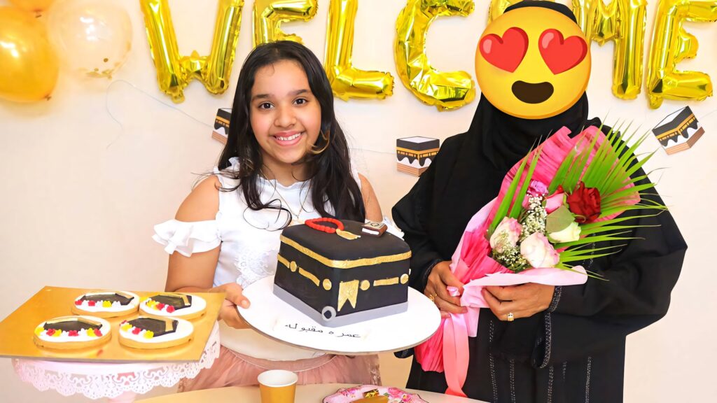 Shfa smiling with Kaaba cake festive balloons golden welcome lettering vibrant celebration wallpaper
