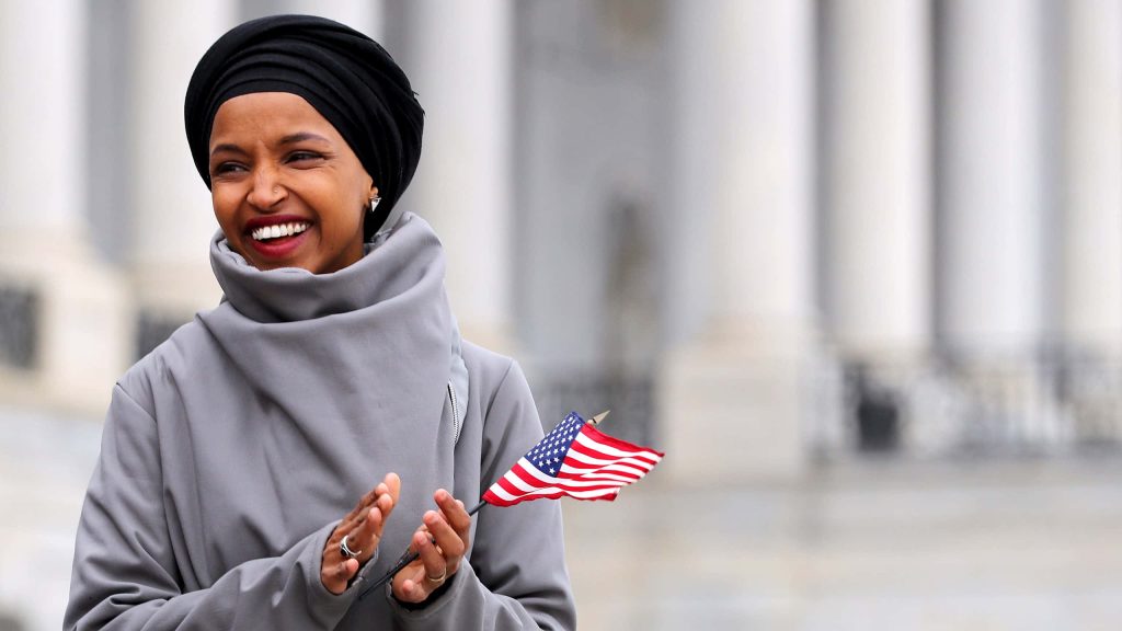 U.S. Representative Ilhan Omar smiling clapping with American flag in bright patriotic setting Wallpaper