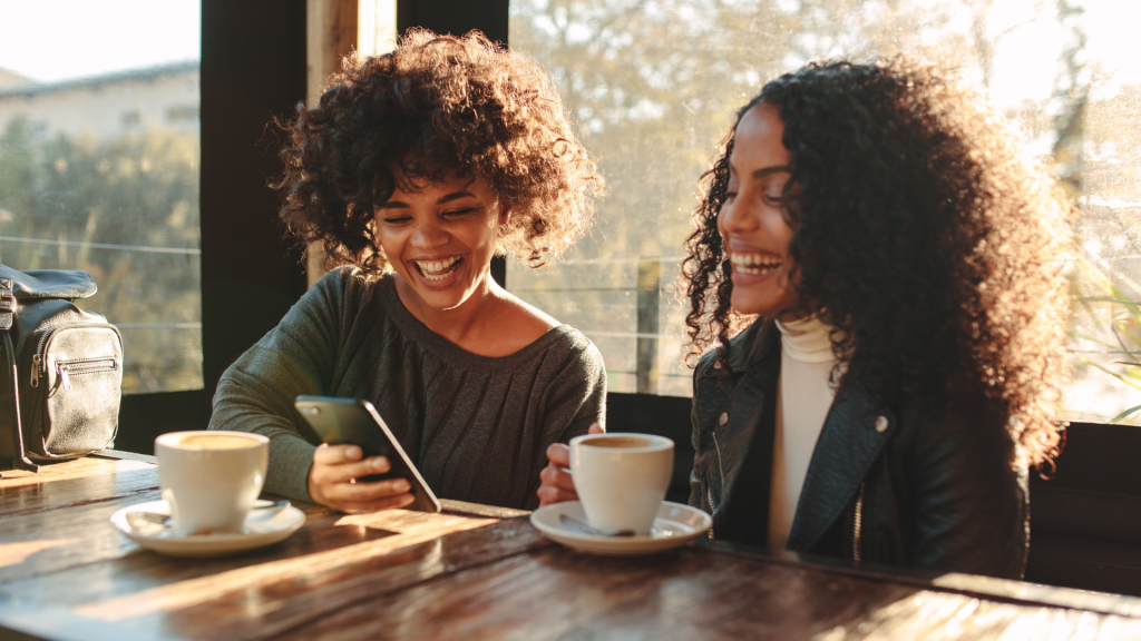 Two joyful women with curly hair laugh over smartphone at coffee shop with warm sunset light Wallpaper
