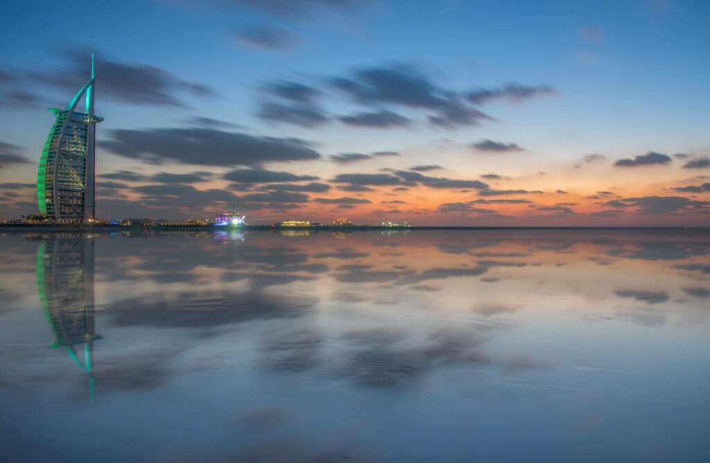 Burj Al Arab sail-shaped hotel reflected on calm water at vibrant sunset with blue and orange hues Wallpaper