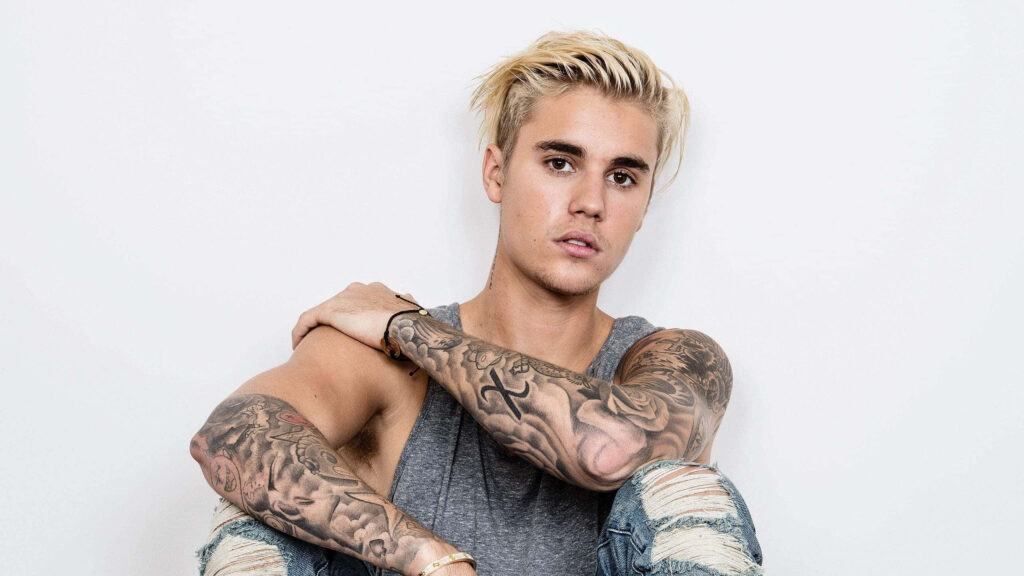 Justin Bieber in sleeveless grey shirt ripped jeans showcasing detailed arm tattoos on white background Wallpaper