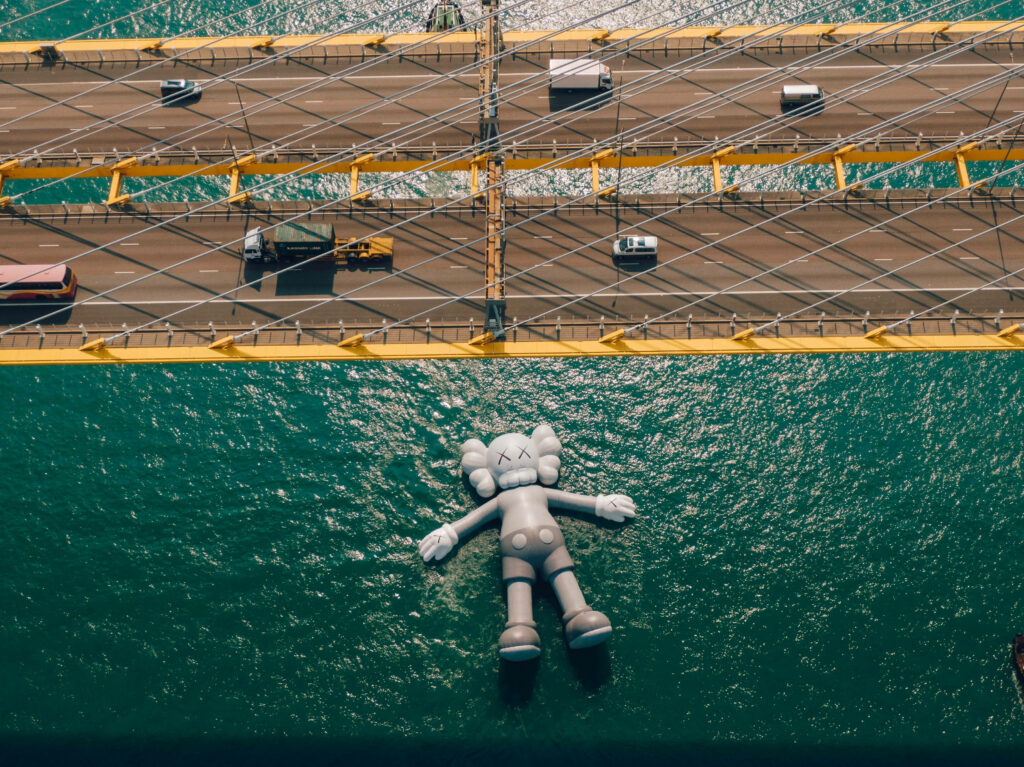 Large KAWS floating sculpture under busy bridge modern art scene aerial view wallpaper background