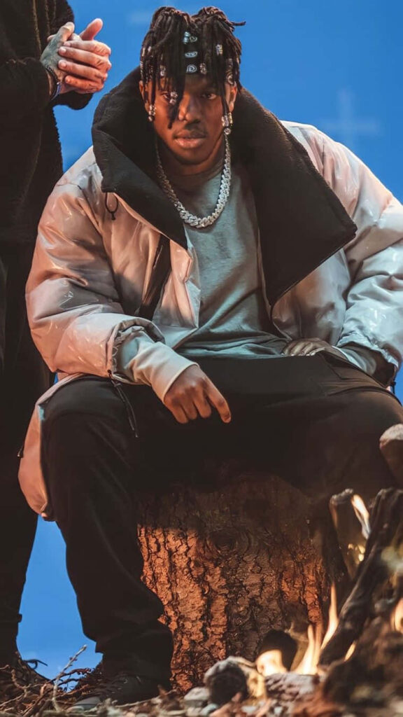 KSI British influencer boxer musician sits confidently by campfire with vibrant blue wallpaper background