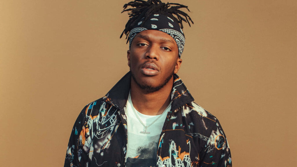 KSI wearing patterned jacket and bandana poses confidently against warm brown wallpaper background