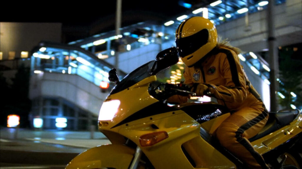 Kill Bill rider in bright yellow suit on matching motorcycle speeding through city wallpaper