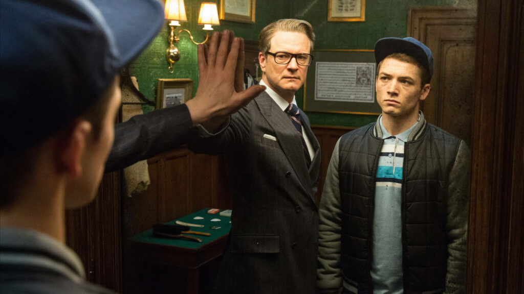 Sharp-dressed Kingsman agent and young recruit in refined room serious moment wallpaper