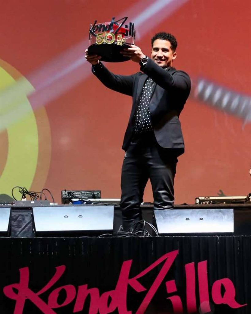 KondZilla in black suit holding 50 Milhões trophy on stage with glowing red backdrop celebrating milestone Wallpaper
