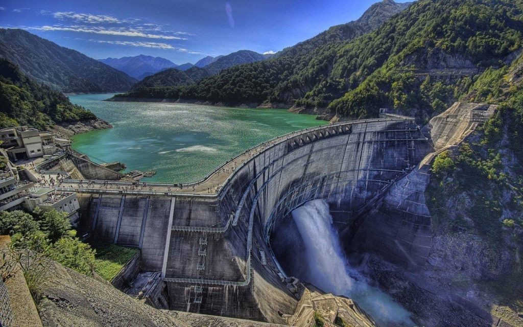Kurobe Dam high angle view releasing water in lush green mountains under clear blue sky Wallpaper