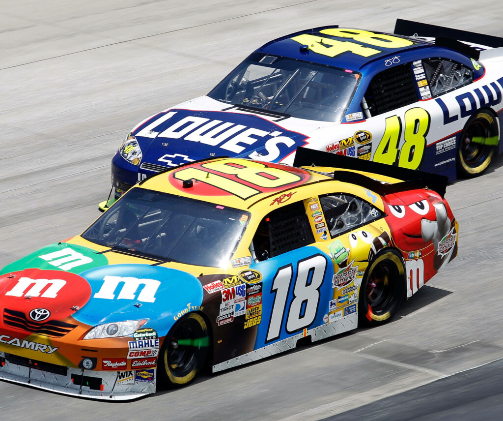 Kyle Busch colorful M&M’s race car overtakes Lowe’s car on NASCAR track in thrilling competition Wallpaper