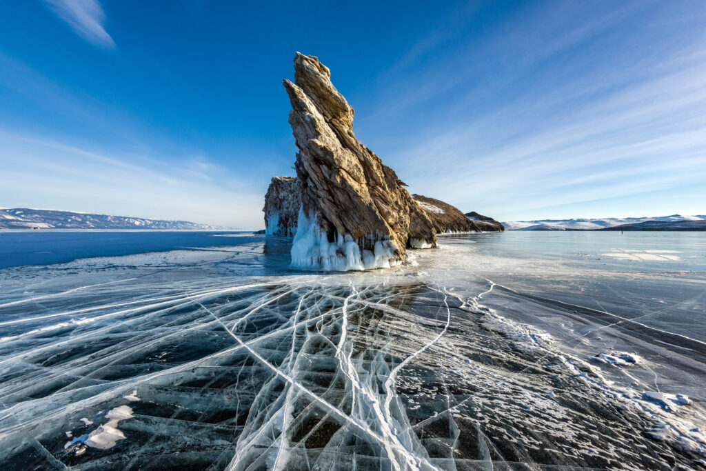 Jagged rock formation rises from frozen Lake Baikal with intricate ice cracks under blue sky Wallpaper