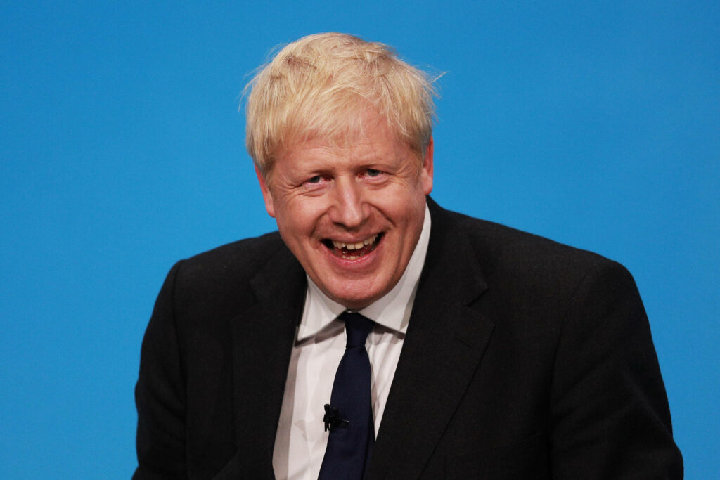 Laughing Boris Johnson wearing dark suit and tie with blonde hair on blue wallpaper background