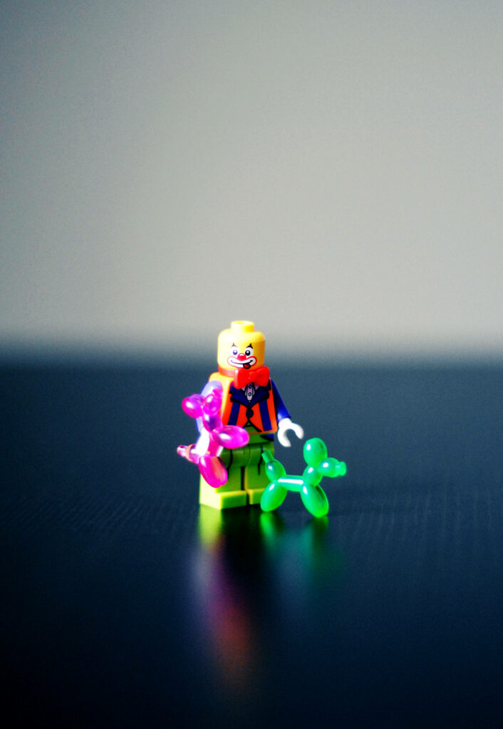 Tiny Lego Ninjago clown figure holding colorful balloon animals on dark surface background Wallpaper