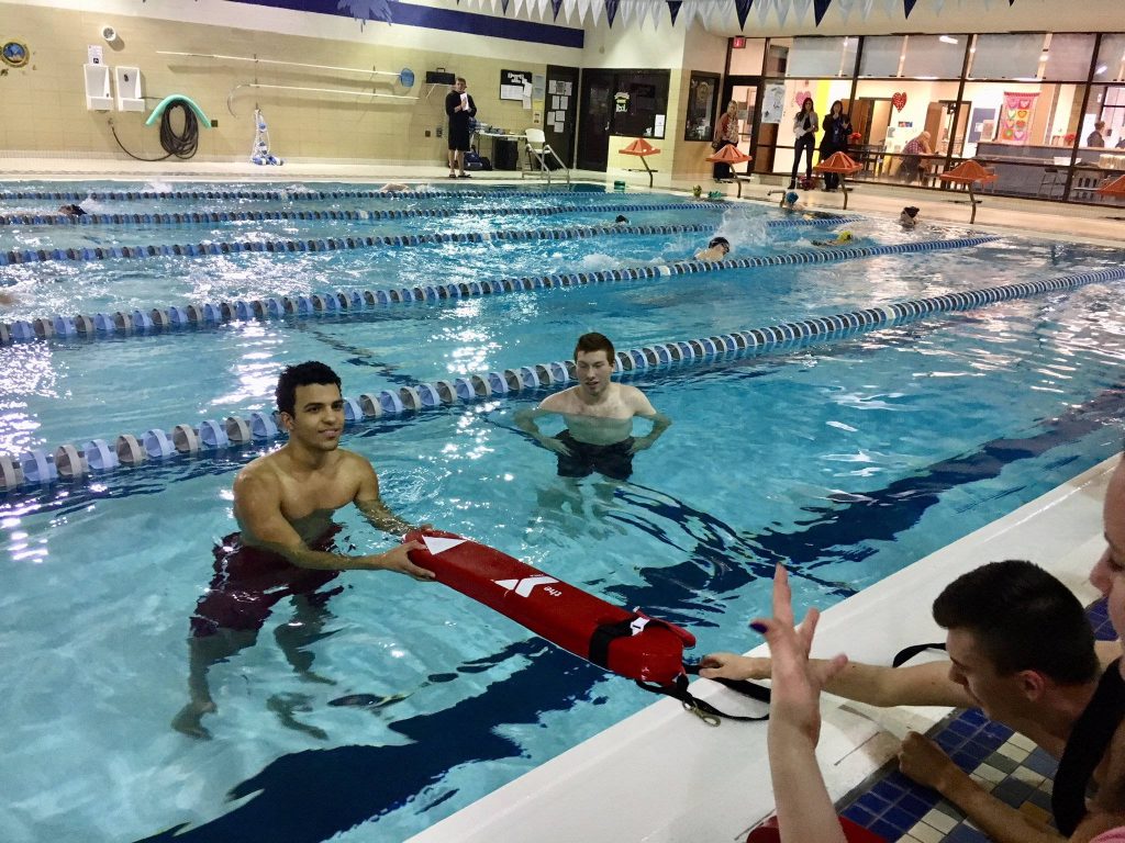 Lifeguarding class with two men practicing water rescue using red YMCA tube in pool Wallpaper