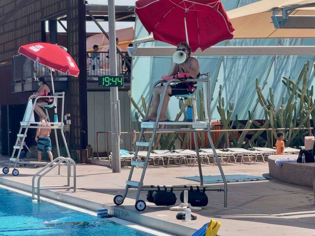 Lifeguards on tall chairs with red umbrellas at sunny desert poolside busy day background Wallpaper