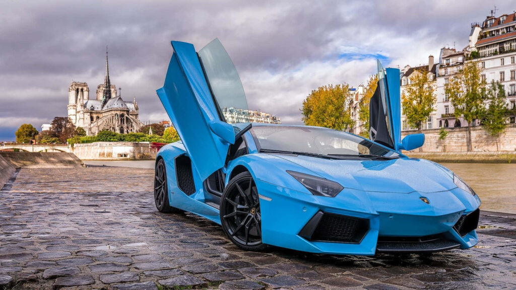 Light blue Lamborghini Aventador with open scissor doors on riverside cobblestone under dramatic sky Wallpaper