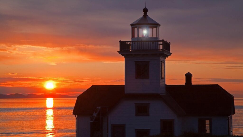 Lighthouse beacon shining at sunset over calm sea with warm orange red hues wallpaper background