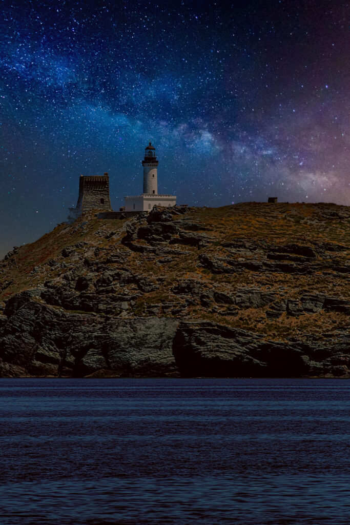 Lighthouse on rocky cliffs at night under Milky Way sky over calm sea tranquil coastal scene Wallpaper