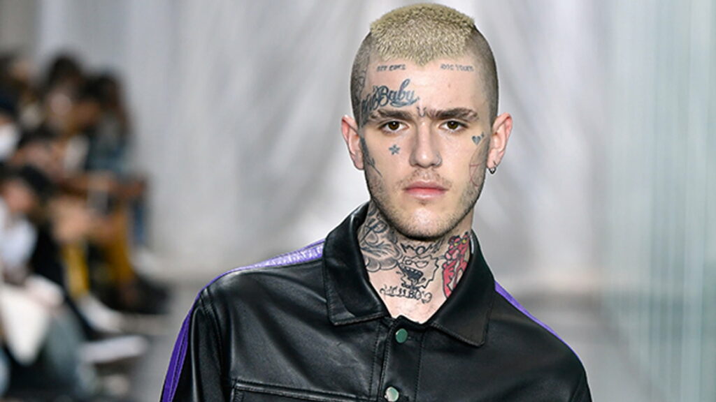 Tattooed Trendsetter: Lil Peep Captivating in Edgy Attire Amidst Blurred Crowd Wallpaper