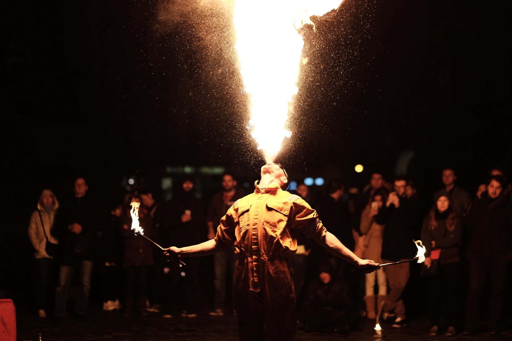 Skilled fire breather in tan jumpsuit exhales large flame at night with glowing torches audience Wallpaper
